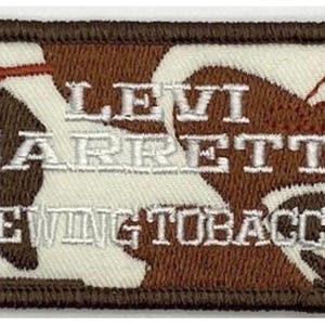 May include: Embroidered patch with a brown border, featuring the text "LEVI GARRETT CHEWING TOBACCO" in white. The patch has a camouflage background in brown, white, and tan, with two orange figures. One figure is holding a rifle, and the other is holding a fishing rod.