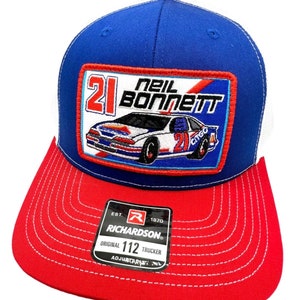 May include: A blue and red trucker hat with a white mesh back. The front of the hat features a patch with a race car and the text "21 Neil Bonnett". The hat also has a Richardson logo and the text "Original 112 Trucker".