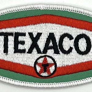 May include: Oval-shaped embroidered patch featuring the Texaco logo. The patch has a green background with a white stitched border. The Texaco logo is in a red and white design with the word "TEXACO" in black.