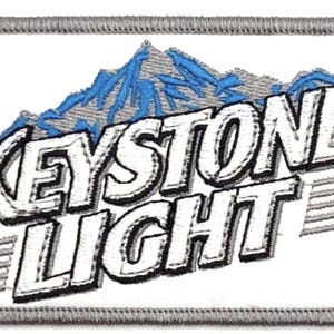 Puede incluir: Parche blanco bordado con una cordillera gris y las palabras "Keystone Light" en blanco con contornos negros.