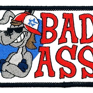 May include: Embroidered patch with a cartoon dog wearing sunglasses and a red baseball cap. The dog has a toothy grin and is standing with its arms crossed. The text "BAD ASS" is in red letters.