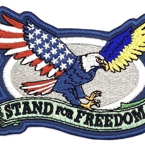 May include: Embroidered patch featuring a blue and yellow eagle with outstretched wings. The eagle's wings are decorated with the American flag and a blue and yellow pattern. The eagle is flying over a white oval with a blue border. Below the eagle is a green banner with the text "Stand for Freedom".