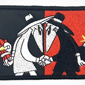 May include: Embroidered patch featuring two cartoon figures shaking hands. The figure on the left is wearing a white suit and holding a lit stick of dynamite. The figure on the right is wearing a black suit and holding a lit bomb. The background is split in half, with black on the left and red on the right.