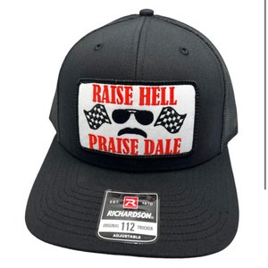 Op de afbeelding: Zwarte truckercap met een rood-witte patch waarop "Raise Hell Praise Dale" staat, met een cartoon gezicht met een zonnebril en een snor. De pet is verstelbaar en heeft een Richardson-logo.