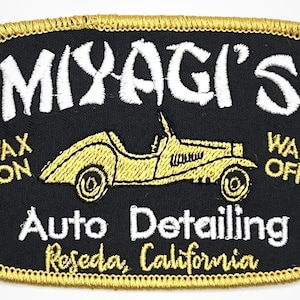 Peut inclure: Écusson brodé avec un fond noir et une bordure dorée. L'écusson présente le texte "Miyagi's" en blanc, une voiture vintage jaune et les mots "Wax On" et "Wax Off". Le texte "Auto Detailing, Reseda, California" est également inclus.