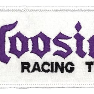 Op de afbeelding: Witte rechthoekige patch met paarse geborduurde tekst "Hoosier" en zwarte tekst "Racing Tire" met een geregistreerd handelsmerk.