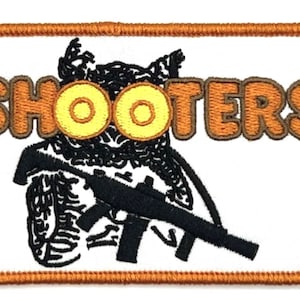 May include: An embroidered patch with a brown outline and white background. The patch features a cartoon owl wearing a pair of yellow goggles and holding a rifle. The text "SHOOTERS" is written in brown letters above the owl.