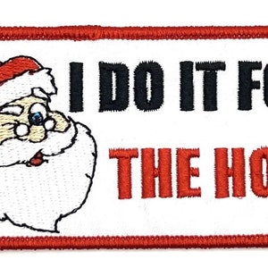 Könnte beinhalten: Ein rechteckiger weißer Patch mit rotem Rand. Der Patch zeigt ein gesticktes Bild vom Weihnachtsmann mit einem zwinkernden Auge und dem Text "I DO IT FOR THE HO'S" in schwarzer und roter Schrift.
