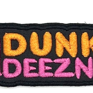 Op de afbeelding: Een zwarte geborduurde patch met de tekst "Dunkin' Deeznuts" in oranje en roze letters. Een gestileerd beeld van een koffiebeker met stoom die uit de bovenkant komt, bevindt zich aan de linkerkant van de patch.