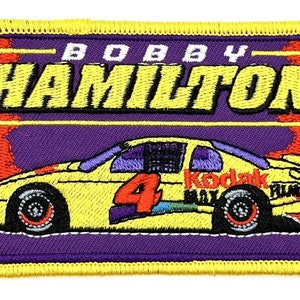 Puede incluir: Parche bordado amarillo con un coche de carreras amarillo con el número 4 en el lateral. El coche tiene un parabrisas morado y un logotipo de Kodak rojo y blanco en el lateral. El texto "BOBBY HAMILTON" está por encima del coche en amarillo.