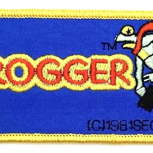 May include: A blue embroidered patch with the word "FROGGER" in yellow and red letters. A cartoon frog wearing a red headband is jumping to the right, holding a black object. The text "(C)1981 SEGA" is in the bottom right corner.