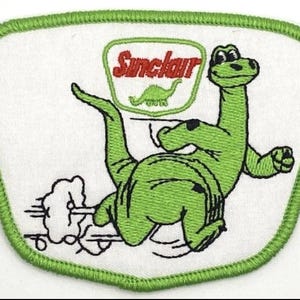 May include: Embroidered patch featuring a green dinosaur with the Sinclair logo. The dinosaur is depicted running with a cloud of smoke behind it. The patch has a white background with a green border.