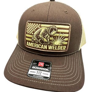 May include: A brown and beige trucker cap with a patch showing an American flag and a welder. The patch reads "AMERICAN WELDER" in gold lettering. The cap is adjustable and has a Richardson 112 tag.