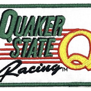 Könnte beinhalten: Gestickter Aufnäher mit dem Schriftzug "Quaker State Racing" in Grün und Schwarz. Das Design umfasst ein großes gelbes "Q" und rote horizontale Linien, alles vor einem weißen Hintergrund mit grünem Rand.