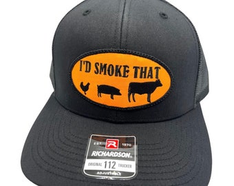 May include: Black trucker hat with an orange oval patch that says "I'd Smoke That" with a chicken, pig, and cow silhouette. The hat has a black mesh back and a Richardson 112 label.