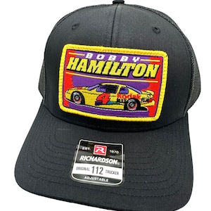 Puede incluir: Gorra de camionero negra con un parche que presenta "Bobby Hamilton" y un coche de carreras amarillo con el número 4. El parche tiene detalles en rojo, morado y amarillo. La gorra tiene una visera curva y una parte trasera de malla. La etiqueta dice "Richardson Original 112 Trucker".