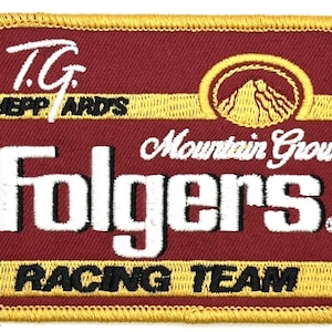 Puede incluir: Parche bordado con fondo granate y ribete dorado. El parche presenta las palabras "T.G. Sheppard's" y "Folgers" en blanco, con "Mountain Grown" en cursiva. La parte inferior dice "Racing Team" en negro.