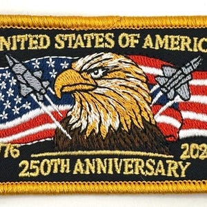 May include: Embroidered patch with a gold border, featuring an eagle, American flag, and text. The text reads "UNITED STATES OF AMERICA", "1776", "2026", and "250TH ANNIVERSARY". The eagle is brown, white, and yellow.