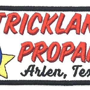 May include: A white embroidered patch with a black border. The patch features a red star with a blue flame in the centre. The text "STRICKLAND PROPANE" is in red and the text "Arlen, Texas" is in black.