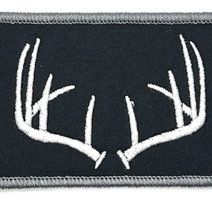 May include: A rectangular patch with a black background and a white embroidered antler design. The patch has a gray border and is likely for clothing or accessories.