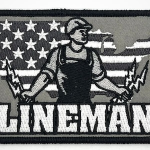 May include: A gray embroidered patch with a white outline. The patch features a cartoon lineman holding electrical wires in front of a stylized American flag. The text "LINEMAN" is embroidered in white below the image.