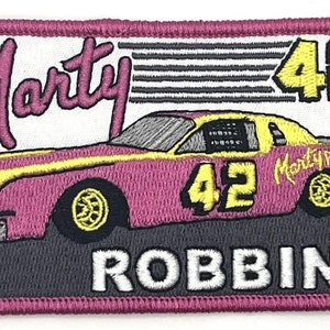 Könnte beinhalten: Gestickter Aufnäher mit einem rosa Rennwagen mit gelben Akzenten und der Nummer 42. Der Aufnäher enthält den Namen "Marty Robbins" und einen rosa Rand. Der Hintergrund ist weiß und grau.