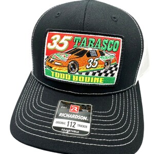 May include: Black and white trucker hat with a red, white, and green embroidered patch featuring a race car with the number 35 and the text "TABASCO" and "TODD BODINE". The hat has a white mesh back and a black front with a white stitched border. The hat also has a black and red label with the text "RICHARDSON" and "ORIGINAL 112 TRUCKER ADJUSTABLE".