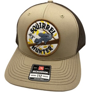 May include: A tan and brown trucker hat with a brown mesh back. The front of the hat features an embroidered patch with a squirrel and the words "Squirrel Hunter". The hat also has a brown leather adjustable strap with a black label that reads "Richardson 112 Original Trucker Adjustable".