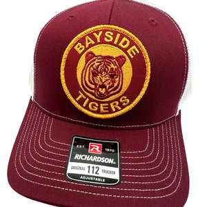 May include: Burgundy and white trucker hat with a gold embroidered patch that says "Bayside Tigers" with a tiger head inside the circle. The hat has a white mesh back and a Richardson 112 adjustable strap.