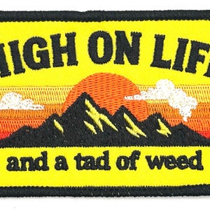 May include: Embroidered patch with a yellow background and black border. The patch features the text "HIGH ON LIFE" above a mountain range with a setting sun and the text "and a tad of weed".
