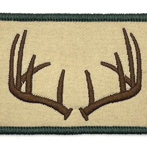 Może przedstawiać: Brązowa naszywka z haftem przedstawiającym parę jelenich rogów na beżowym tle. Naszywka ma ciemnozielony brzeg.