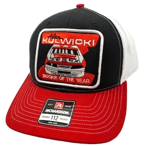 Pode incluir: Um boné de caminhoneiro preto e vermelho com uma malha branca nas costas. O boné apresenta um grande remendo bordado com o nome "Alan Kulwicki" e um carro de corrida com o número "7" nele. O remendo também diz "Thunderbird Rookie of the Year". O boné tem um logotipo Richardson e diz "Original 112 Trucker One Size".