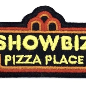 May include: Embroidered patch with the text "SHOWBIZ PIZZA PLACE" in yellow, orange, and black. The patch has a black border and a decorative orange and brown frame. A retro-style design.