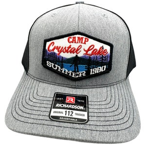 May include: A grey and black trucker hat with a black mesh back. The front of the hat features a black and white embroidered patch that says "Camp Crystal Lake Summer 1980". The patch has a blue lake, green trees, and a silhouette of a person holding an axe. The hat also has a black and white label that says "Richardson 112 Original Trucker Adjustable".