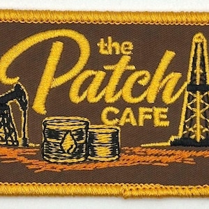 May include: Embroidered patch with the words "the Patch CAFE" in yellow script and block letters. The patch features images of an oil derrick, oil barrels, and a pump jack, all in black and yellow on a brown background.