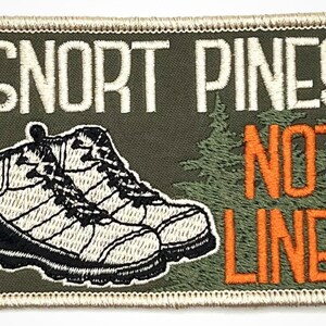 May include: An embroidered patch in green with white text that reads "Snort Pines Not Lines" and a pair of brown hiking boots. The patch has a white border.