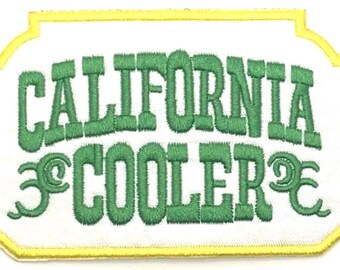 Op de afbeelding: Een witte geborduurde patch met een gele rand. De patch heeft de tekst "CALIFORNIA COOLER" in groene letters met een gestileerde "C" aan weerszijden van het woord "COOLER".