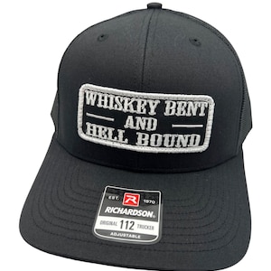 Könnte beinhalten: Schwarze Trucker-Kappe mit einem weißen Patch, auf dem "Whiskey Bent and Hell Bound" steht. Die Kappe ist verstellbar und hat ein Richardson 112 Logo an der Seite.