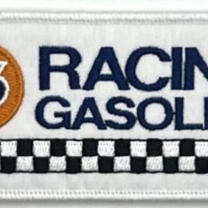 Pode incluir: Patch bordado com o texto "RACING GASOLINE" em azul marinho, um logótipo circular com o número "76" e um padrão xadrez a preto e branco. O patch é retangular com fundo e borda brancos.