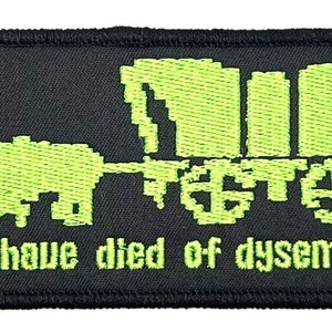 Può includere: Una toppa ricamata nera con un'immagine pixelata verde di un carro coperto trainato da un bue. Il testo "You have died of dysentery." è sotto l'immagine.