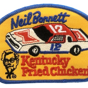 Op de afbeelding: Een geborduurde patch in geel en blauw met een witte raceauto met rode en blauwe accenten. De auto heeft het nummer "12" aan de zijkant en de woorden "Neil Bonnett" en "Kentucky Fried Chicken" zijn geborduurd op de patch.