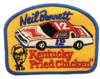 May include: A yellow and blue embroidered patch featuring a white race car with red and blue accents. The car has the number "12" on the side and the words "Neil Bonnett" and "Kentucky Fried Chicken" are embroidered on the patch.