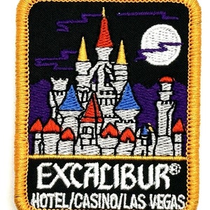 Puede incluir: Un parche bordado negro con borde dorado que presenta un castillo blanco con detalles rojos, azules y amarillos. El castillo está situado contra un cielo nocturno morado y negro con una luna blanca. El texto "EXCALIBUR® HOTEL/CASINO/LAS VEGAS" está impreso en blanco debajo del castillo.