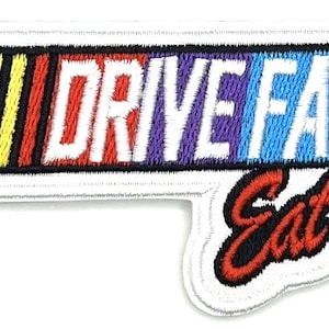 Könnte beinhalten: Gestickter Aufnäher mit den Worten "DRIVE FAST" in weißen Großbuchstaben, über den Worten "Eat ass" in roter Schrift. Der Aufnäher hat ein Rennstreifen-Design mit schwarzen, gelben und roten Streifen.