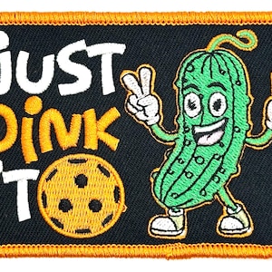 May include: Embroidered patch with a black background and an orange border. The patch features a cartoon pickle character giving a peace sign. Text reads "Just Dink It" in white and yellow.