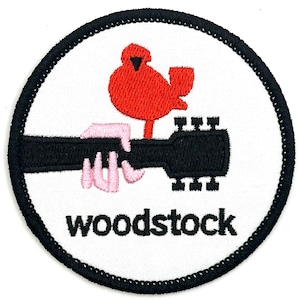 Puede incluir: Un parche circular blanco con un borde negro. El parche presenta un pájaro rojo posado en una guitarra negra con una mano rosa sosteniendo el cuello. El texto "woodstock" está escrito en negro debajo de la guitarra.