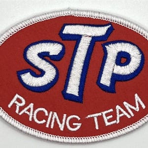 Puede incluir: Parche ovalado del equipo de carreras STP. El parche presenta las letras "STP" en blanco con contornos azules, sobre un fondo rojo. Las palabras "RACING TEAM" están bordadas en blanco a lo largo del borde inferior.