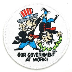 Puede incluir: Parche blanco bordado con una imagen de dibujos animados del Tío Sam sosteniendo a un hombre boca abajo por el cuello. El hombre lleva traje y corbata y tiene dinero que se le cae de los bolsillos. El texto "OUR GOVERNMENT AT WORK!" está impreso debajo de la imagen.