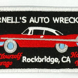 Op de afbeelding: Een geborduurde zwarte en witte patch met een rode auto en de tekst "Darnell's Auto Wrecking", "Do It Yourself Garage", "Rockbridge, CA" en "Honk for Entry".