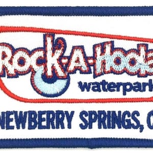 May include: A rectangular embroidered patch with a white background and a dark blue border. It features the red-outlined text Rock-A-Hoola waterpark and the location NEWBERRY SPRINGS, CA in dark blue, with a light blue circular graphic.